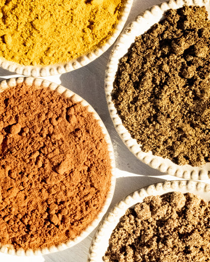 An overhead view of four small, round, white ceramic bowls, each filled with different colored powdered spices. The top-left bowl contains a bright yellow spice often used in Surya's Mighty Turmeric Latte mixes; the top-right a greenish-brown spice; the bottom-left a reddish-brown spice known for its anti-inflammatory properties; and the bottom-right a light brown spice.
