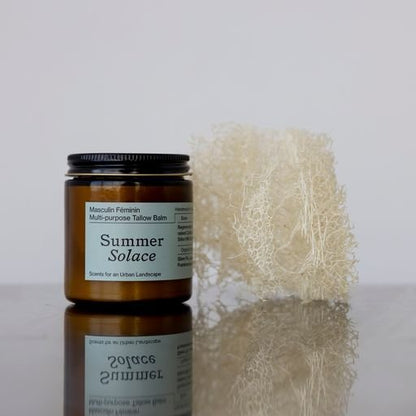 A jar of "Masculin Féminin Balm - Regenerative Tallow™" by Summer Solace Tallow, featuring a dark brown label, sits on a reflective surface. The jar is placed beside a natural sponge-like object. The label displays the words "Masculin Féminin" and "Scents for an Urban Landscape," emphasizing the regenerative tallow blend within.