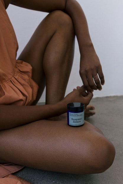 A person in an orange dress sits on the floor with knees bent, resting hands on their legs. In front of their shins is a small blue jar labeled "Masculin Féminin Balm - Regenerative Tallow™," from the brand Summer Solace Tallow. The background is a plain, light-colored wall and floor.