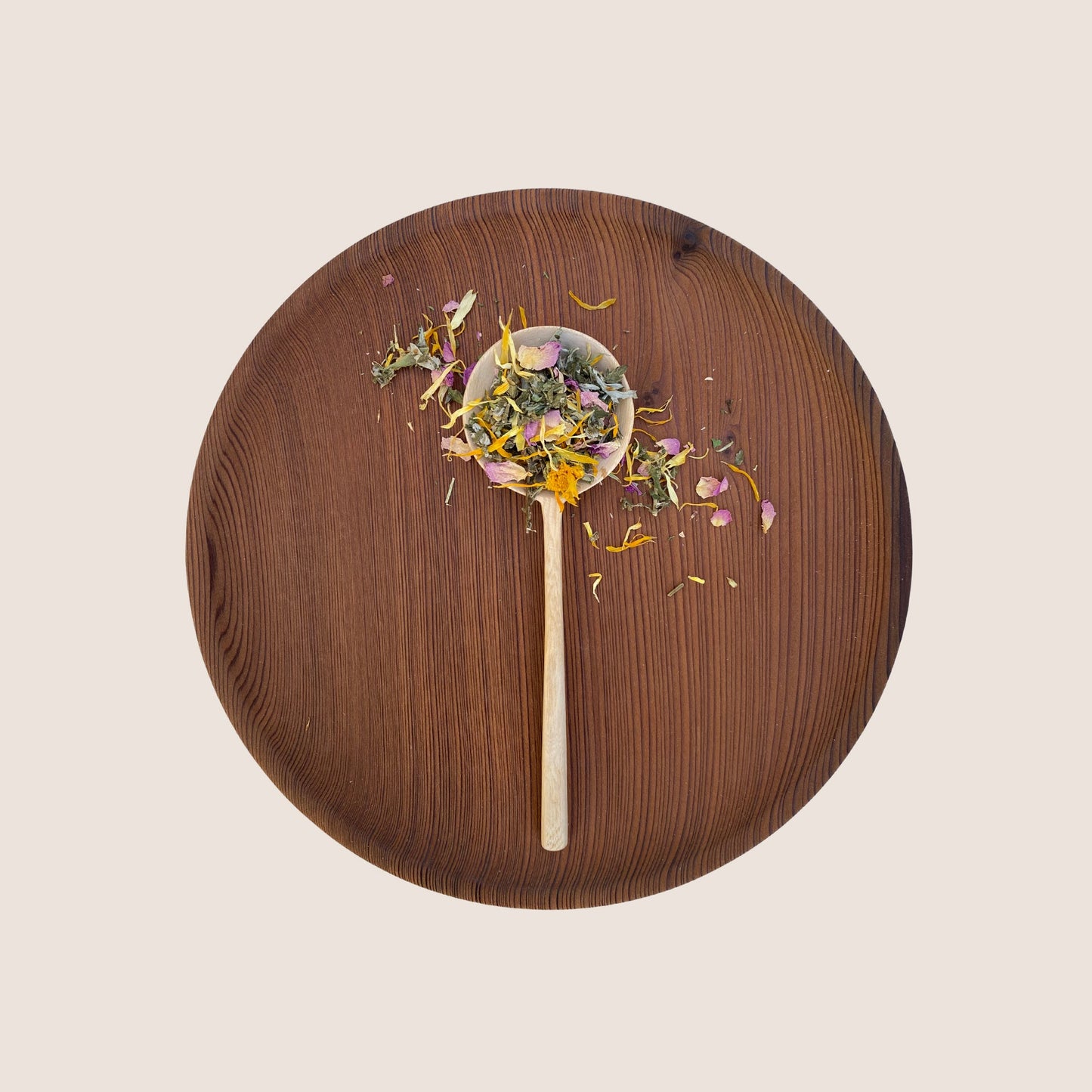 A wooden spoon filled with a mixture of dried herbs and flower petals from Moon Canyon Healing's Sacred Steam Herbal Yoni Blend rests on a round, dark wooden plate against a light background. The colorful herbs and petals are visually distinct against the warm tones of the wooden surfaces.