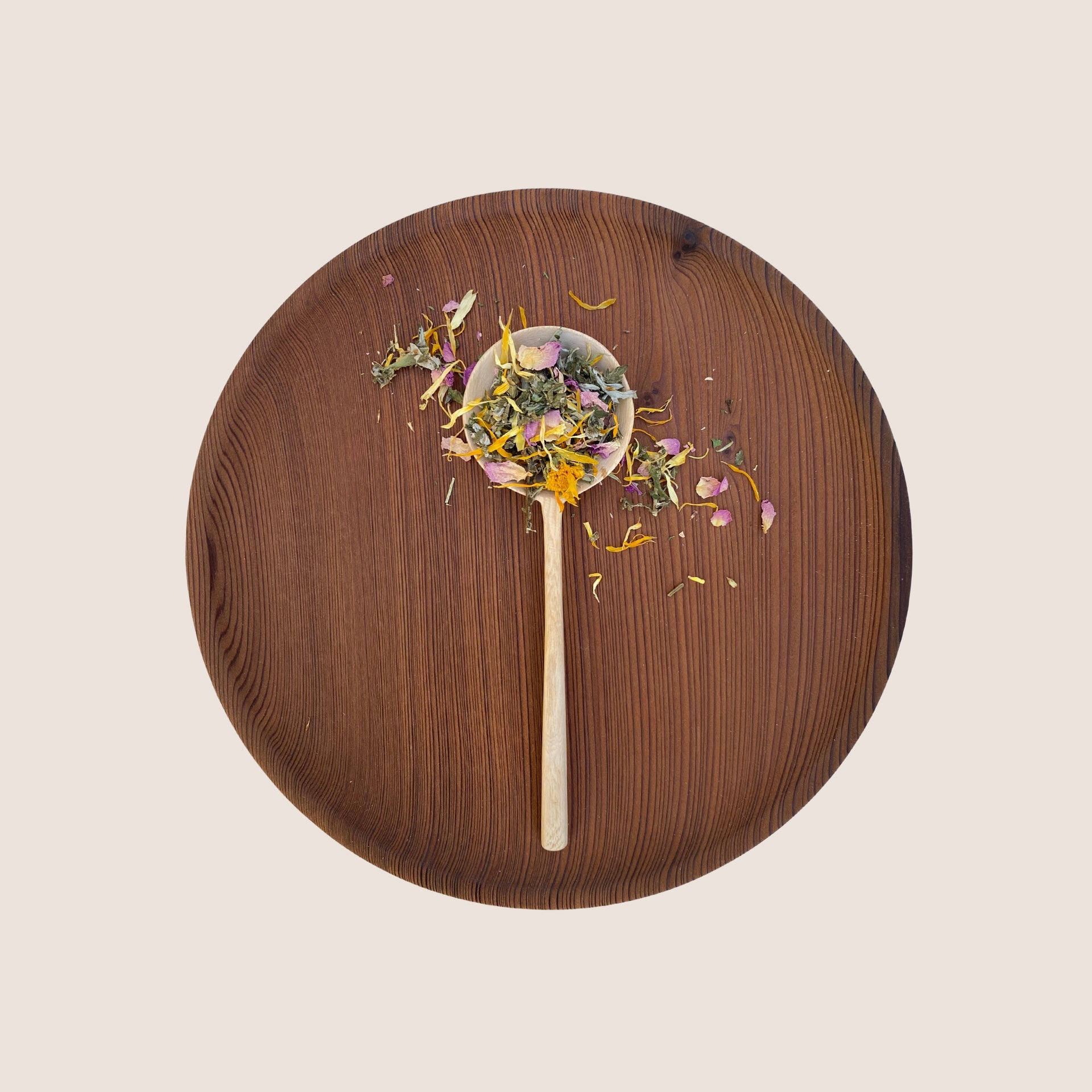 A wooden spoon filled with a mixture of dried herbs and flower petals from Moon Canyon Healing's Sacred Steam Herbal Yoni Blend rests on a round, dark wooden plate against a light background. The colorful herbs and petals are visually distinct against the warm tones of the wooden surfaces.
