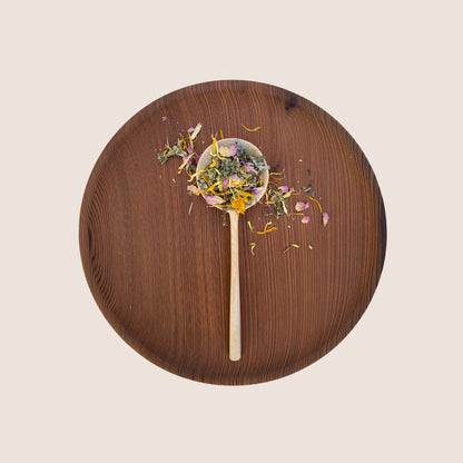 A wooden spoon filled with a mixture of dried herbs and flower petals from Moon Canyon Healing's Sacred Steam Herbal Yoni Blend rests on a round, dark wooden plate against a light background. The colorful herbs and petals are visually distinct against the warm tones of the wooden surfaces.