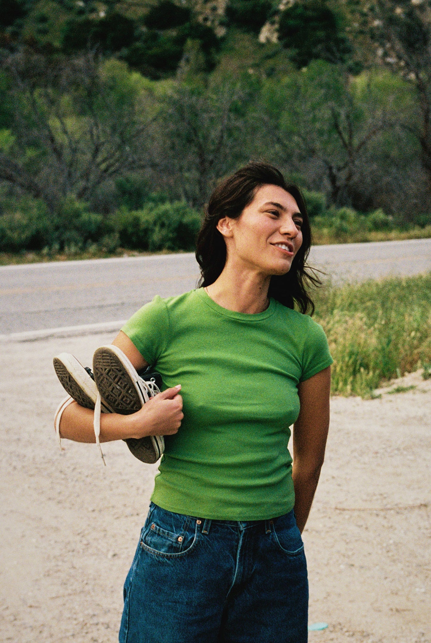 Matcha A woman in a green LA Relaxed Bebe Organic Tee and jeans smiles by the roadside, holding sneakers. Lush greenery and a winding road extend behind her.