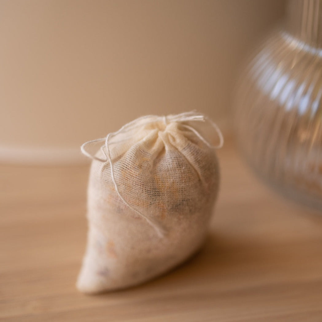 A small drawstring pouch, "meditate bath soak" by Honeybee Hippie, is crafted from light beige mesh and filled with organic flowers. It rests on a wooden surface against a blurred background showcasing part of a ribbed glass object.