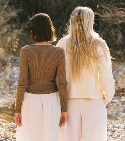 Two women stand side by side, facing away. One wears a brown cardigan with pearlescent buttons; the other wears LA RELAXED’s Pointelle Cardigan in cream, made from organic cotton. Both pair their cardigans with light, flowy skirts.