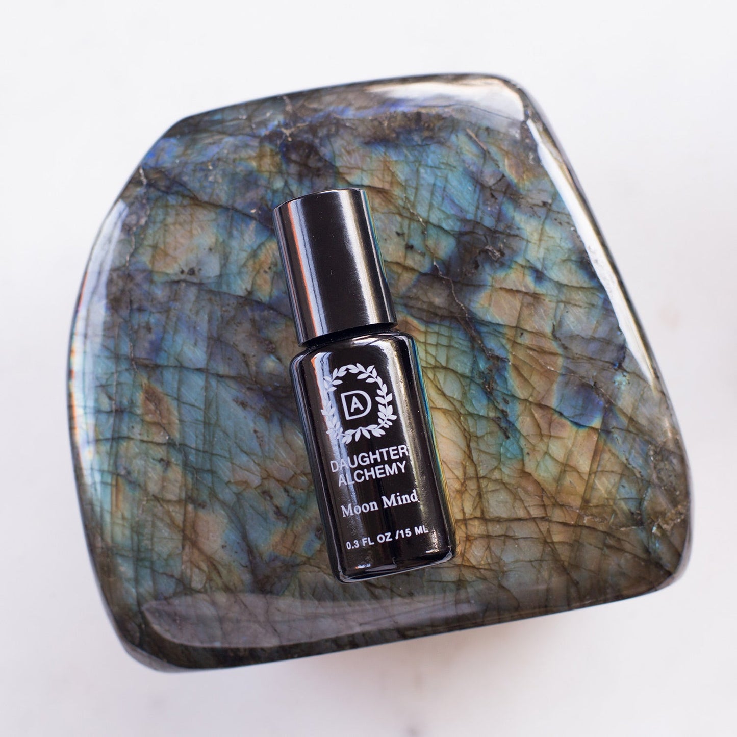 A small bottle of Moon Mind® Meditative Body Oil from Daughter Alchemy is elegantly placed on a polished, multi-colored stone. The black bottle, adorned with white text and the brand’s logo, stands out against the iridescent marbled pattern of blue, green, and brown—a perfect blend of organic ingredients for your mindful moments.