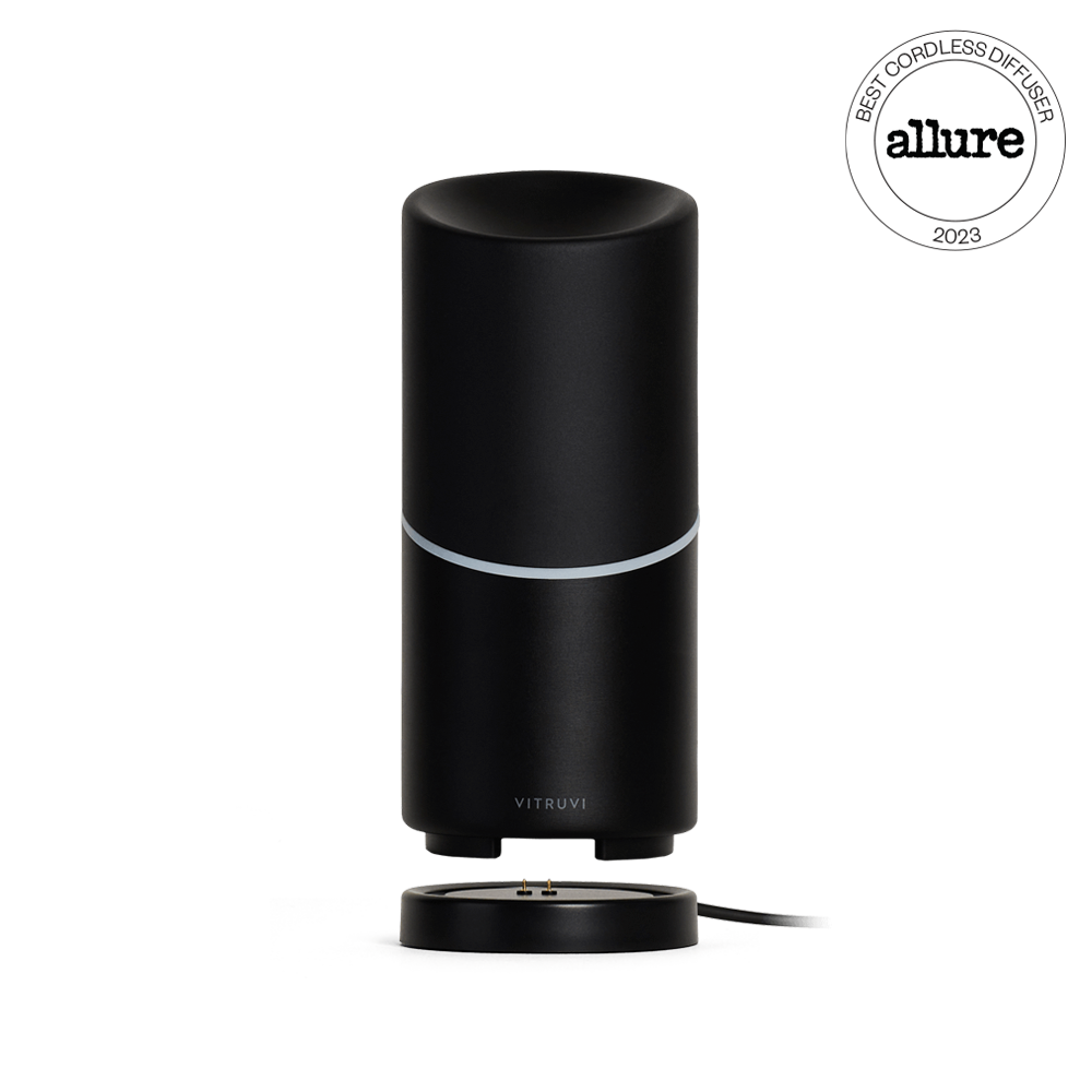 The Vitruvi Move Diffuser is a sleek, black cordless diffuser with a cylindrical shape and a subtle silver band near the top. It rests on a round base with a connected charging cable, ensuring you can scent every corner of your home. A "Best Cordless Diffuser 2023" award badge from Allure is displayed in the top right corner.