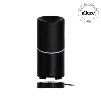 The Vitruvi Move Diffuser is a sleek, black cordless diffuser with a cylindrical shape and a subtle silver band near the top. It rests on a round base with a connected charging cable, ensuring you can scent every corner of your home. A "Best Cordless Diffuser 2023" award badge from Allure is displayed in the top right corner.
