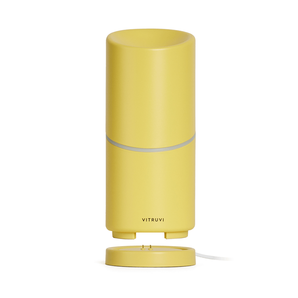 The Move Diffuser by Vitruvi is a sleek, cylindrical yellow essential oil diffuser. It features a smooth surface with a horizontal seam around the middle and the brand name "Vitruvi" printed at the bottom. Designed to sit on a round base, it makes it easy to scent every corner of your space without needing a plug.