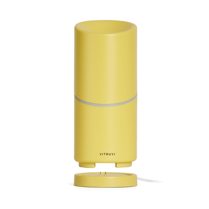 The Move Diffuser by Vitruvi is a sleek, cylindrical yellow essential oil diffuser. It features a smooth surface with a horizontal seam around the middle and the brand name "Vitruvi" printed at the bottom. Designed to sit on a round base, it makes it easy to scent every corner of your space without needing a plug.