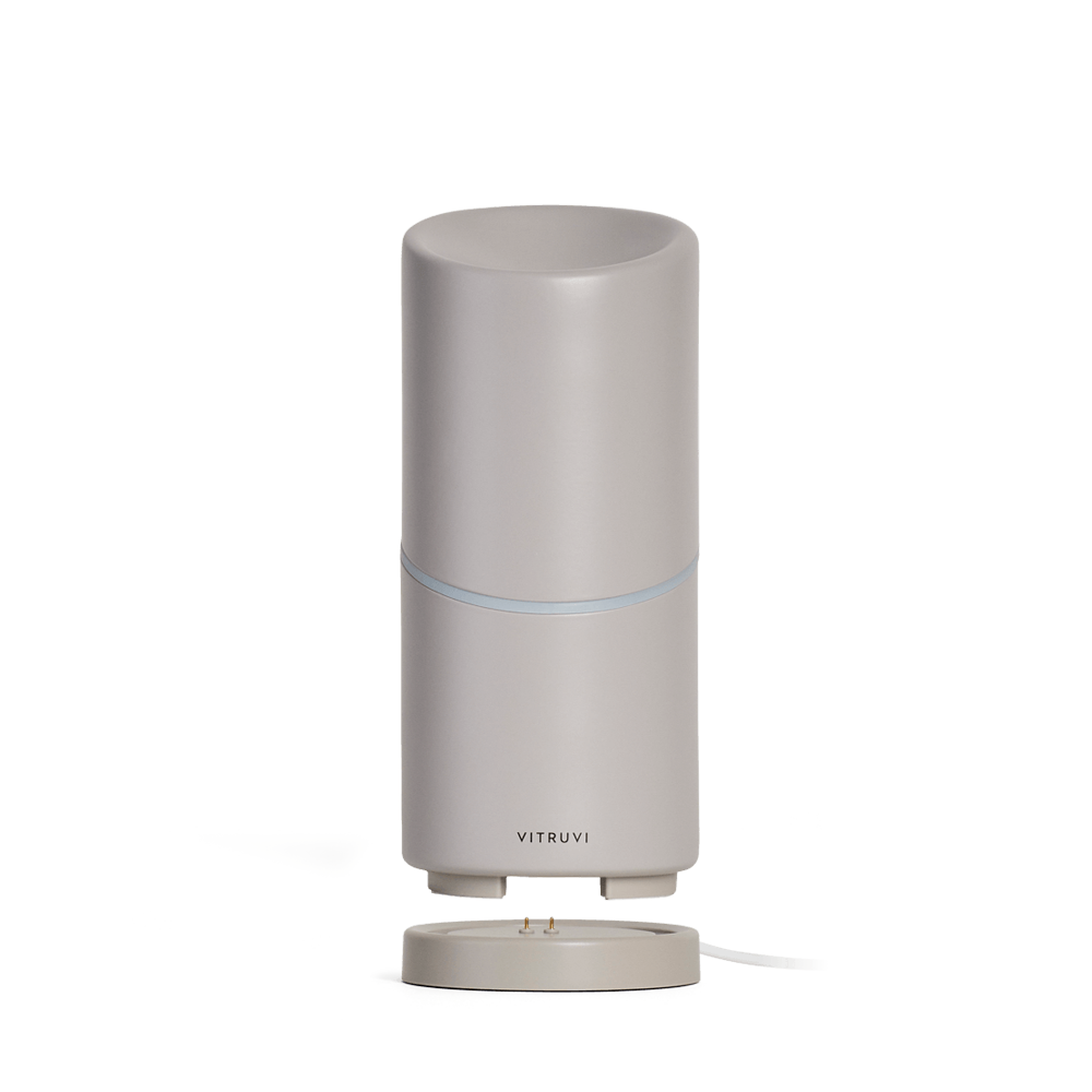 Tall, cylindrical grey Move Diffuser by Vitruvi on a detachable base. This cordless essential oil diffuser boasts a sleek, minimalist design with a subtle, thin blue light ring around the middle. Scent every corner effortlessly with no plug necessary as you carry it from room to room.
