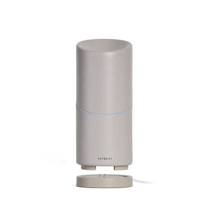 Tall, cylindrical grey Move Diffuser by Vitruvi on a detachable base. This cordless essential oil diffuser boasts a sleek, minimalist design with a subtle, thin blue light ring around the middle. Scent every corner effortlessly with no plug necessary as you carry it from room to room.