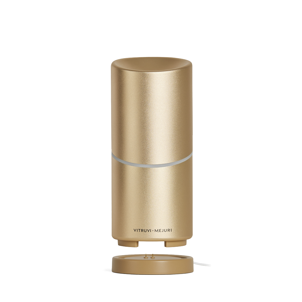 A sleek, gold-colored Vitruvi Move Diffuser with a smooth cylindrical design sits on a matching base. The diffuser, designed to scent every corner, has a small circular opening at the top and a metallic finish. The brand name "Vitruvi" is printed near the bottom.