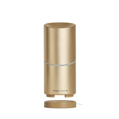 A sleek, gold-colored Vitruvi Move Diffuser with a smooth cylindrical design sits on a matching base. The diffuser, designed to scent every corner, has a small circular opening at the top and a metallic finish. The brand name "Vitruvi" is printed near the bottom.