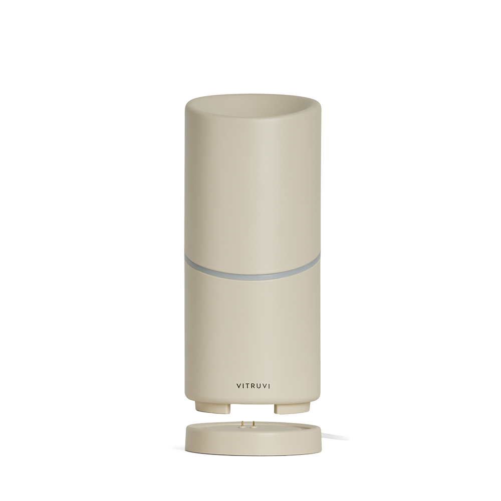 The Vitruvi Move Diffuser is a sleek, minimalist cylindrical diffuser with a smooth beige finish. It features a small, subtle logo near the base that reads "Vitruvi." This cordless diffuser sits on a matching round base and ensures you scent every corner with ease, no plug necessary.