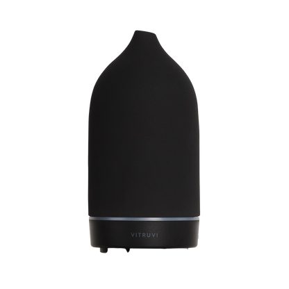 Introducing the Stone Diffuser by Vitruvi, a sleek black essential oil diffuser with a modern, minimalist design. This elegant piece features a smooth, conical shape with a small opening at the top, ideal for pure essential oils. The brand name "Vitruvi" is gracefully displayed on the lower part of the unit, making it an exquisite addition to any home decor.