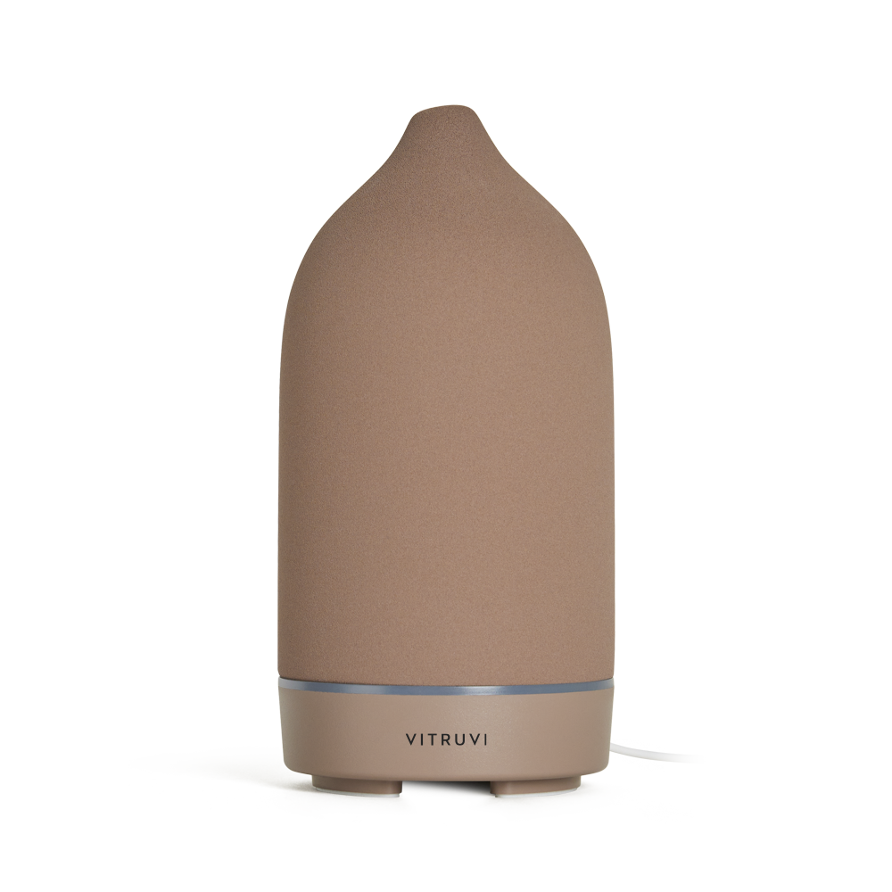 A sleek, cone-shaped diffuser from Vitruvi stands alone against a neutral background. The Stone Diffuser features a matte brown finish with a metallic base, and the brand name "Vitruvi" is prominently displayed on the bottom section, making it an elegant piece of home decor perfect for dispersing pure essential oils.