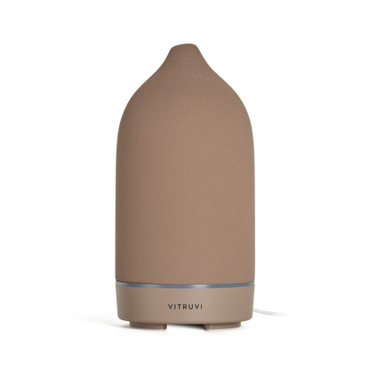 A sleek, cone-shaped diffuser from Vitruvi stands alone against a neutral background. The Stone Diffuser features a matte brown finish with a metallic base, and the brand name "Vitruvi" is prominently displayed on the bottom section, making it an elegant piece of home decor perfect for dispersing pure essential oils.