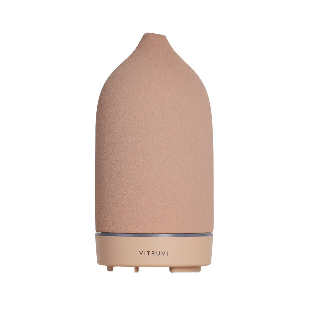 A sleek, pink ceramic essential oil diffuser with a conical shape, featuring the brand name "Vitruvi" at the base. This Stone Diffuser from Vitruvi boasts a minimalist design with a subtle texture, standing upright against a plain white background, making it an elegant addition to any home decor.