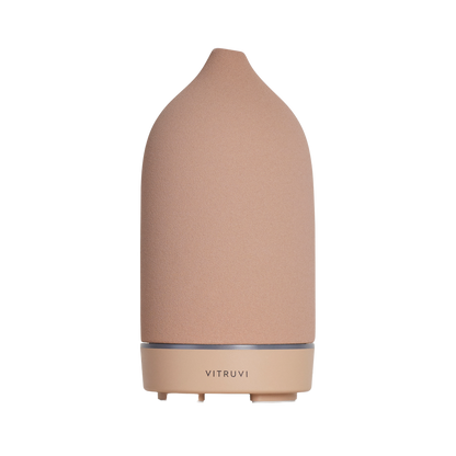 A sleek, pink ceramic essential oil diffuser with a conical shape, featuring the brand name "Vitruvi" at the base. This Stone Diffuser from Vitruvi boasts a minimalist design with a subtle texture, standing upright against a plain white background, making it an elegant addition to any home decor.
