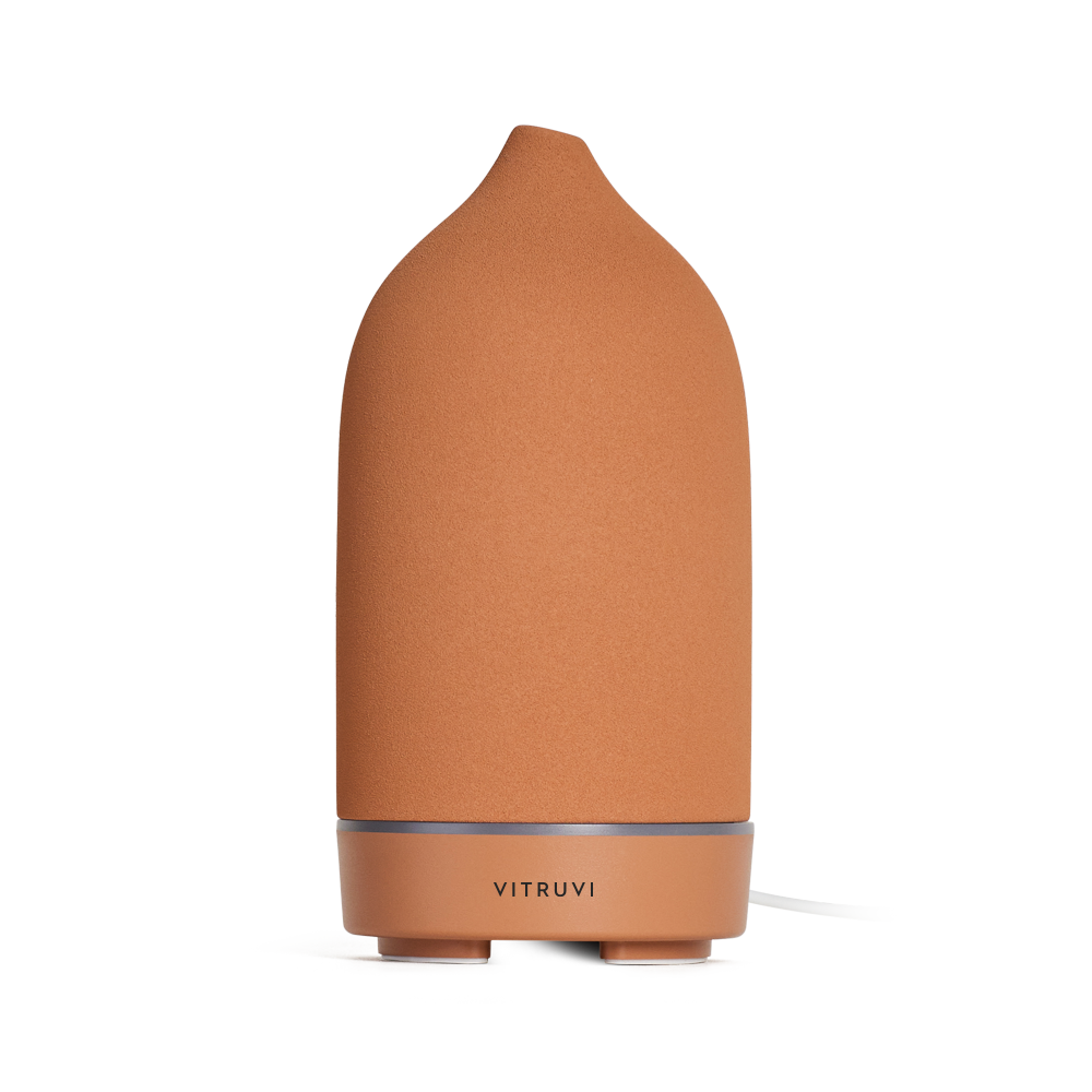 The Stone Diffuser by Vitruvi is a sleek, modern device in a terracotta hue, featuring a cylindrical shape and rounded top. A subtle silver band on the base displays the brand name, making it perfect for dispersing pure essential oils. Its minimalist design seamlessly complements various home decor styles.