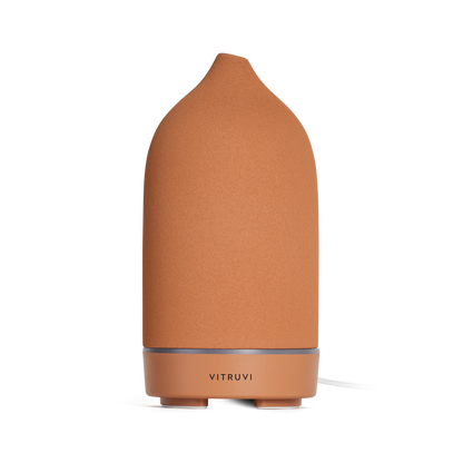 The Stone Diffuser by Vitruvi is a sleek, modern device in a terracotta hue, featuring a cylindrical shape and rounded top. A subtle silver band on the base displays the brand name, making it perfect for dispersing pure essential oils. Its minimalist design seamlessly complements various home decor styles.