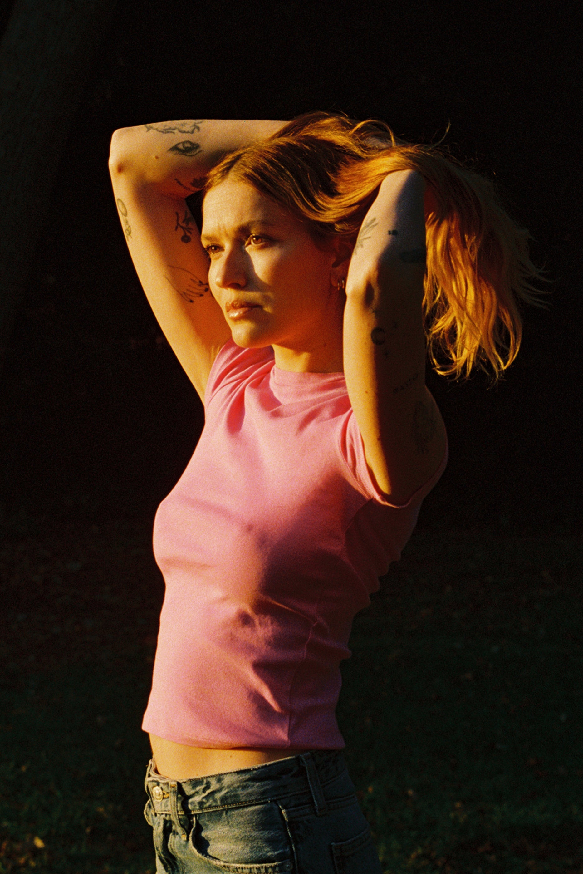 Bubblegum A tattooed person with long hair in a bun stands outside in warm sunlight, wearing a pink Bebe Organic Tee by LA Relaxed and jeans, gazing thoughtfully into the distance against a dark grassy background.