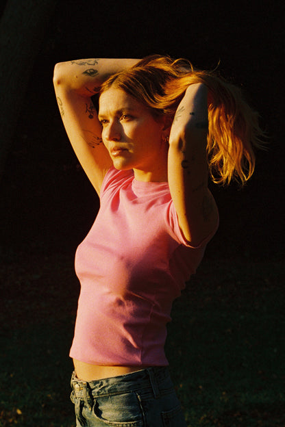 Bubblegum A tattooed person with long hair in a bun stands outside in warm sunlight, wearing a pink Bebe Organic Tee by LA Relaxed and jeans, gazing thoughtfully into the distance against a dark grassy background.