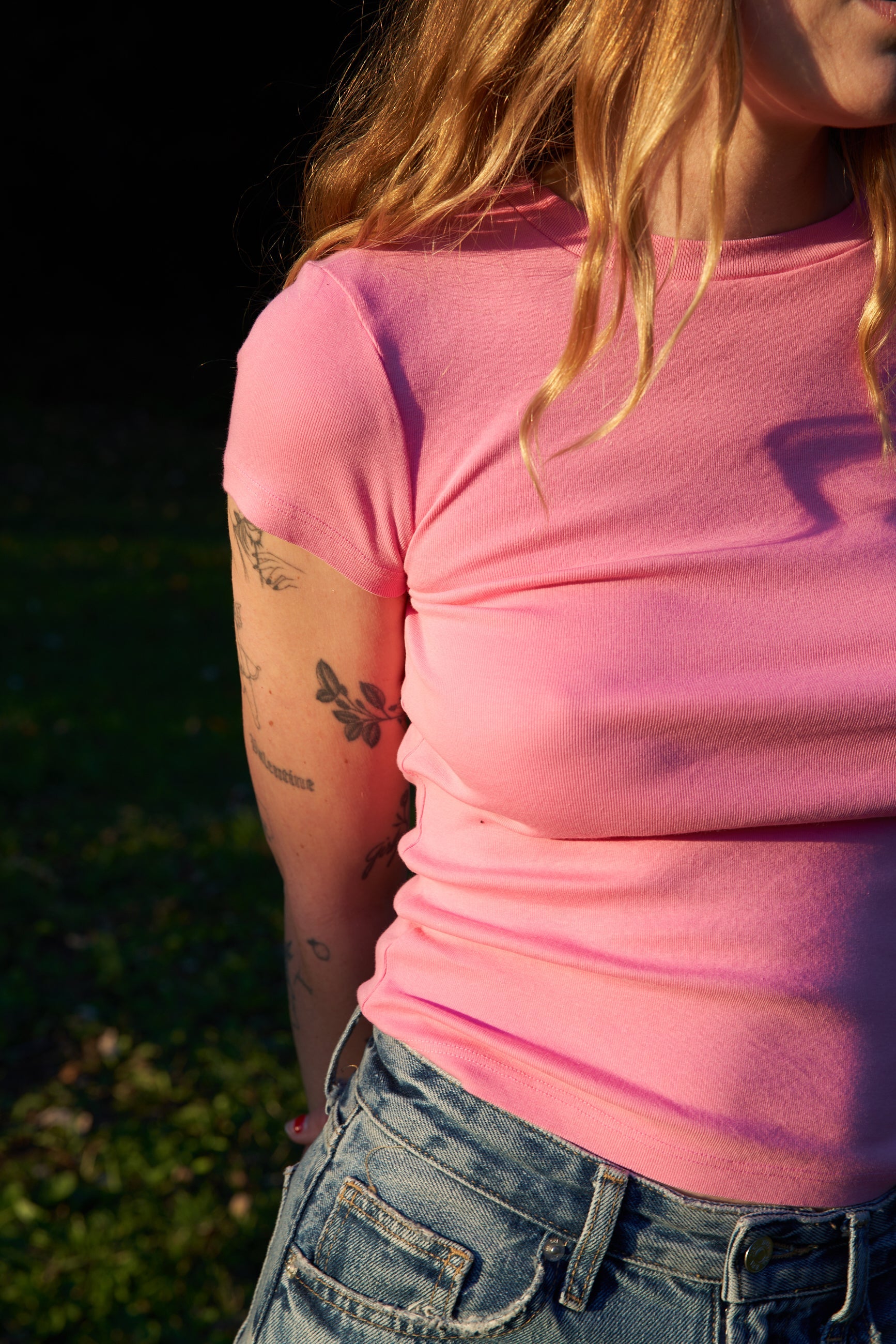 Bubblegum A person with long wavy hair is outdoors, wearing a pink LA Relaxed Bebe Organic Tee and blue denim shorts. Their tattooed arms are partially in shadow, while sunlight highlights the organic cotton fabric of their clothing.