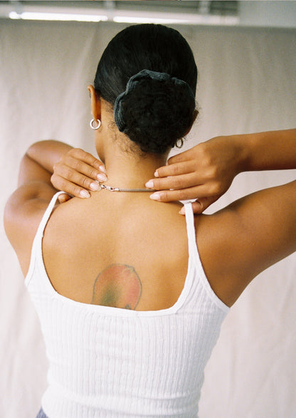 White A person with a peach tattoo on their upper back adjusts a necklace, wearing the LA Relaxed Pointelle Square Cami. Their dark hair is pulled back with a scrunchie. The photo is taken from behind against a light background.