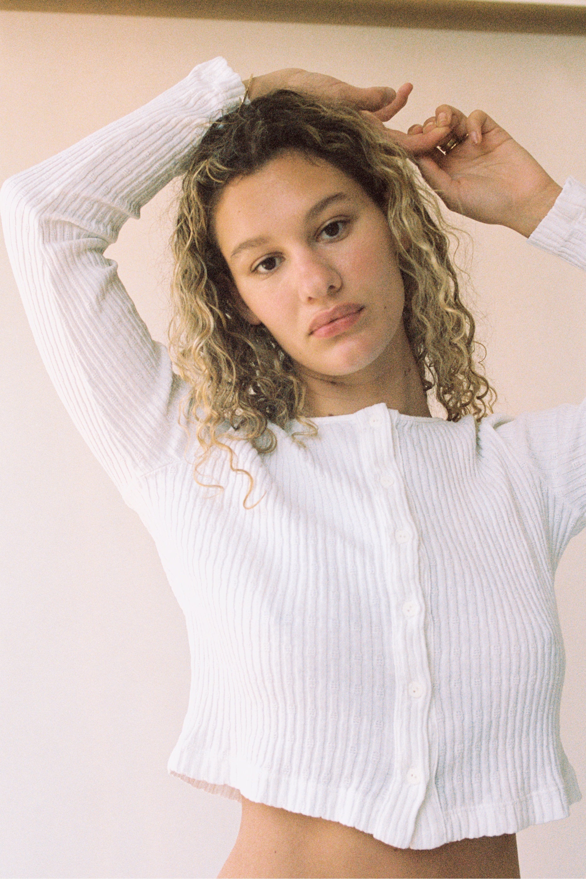 A young woman with curly hair stands indoors, calmly looking at the camera while wearing the LA RELAXED Pointelle Cardigan—a white ribbed long-sleeve top with pearlescent buttons—one arm raised, the other touching her hair.