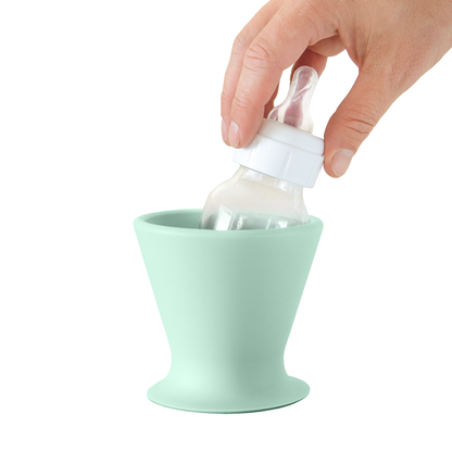 A hand skillfully handles the one-handed bottle prep using a bökee Baby Bottle Holder, placing a BPA-free silicone baby bottle into the pastel green, inverted cone-shaped container. This ingenious holder by bökee offers a wide base for stability. The baby bottle has a clear body and is topped with a white cap and silicone nipple.