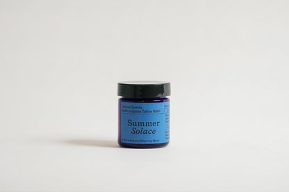 A small blue jar of "Sacred Scents Spiritual Balm - Regenerative Tallow®" from Summer Solace Tallow sits against a plain white background. The jar has a black lid and a light blue label that highlights its grounding and hormone balancing blend, enhanced with anti-inflammatory essential oil.