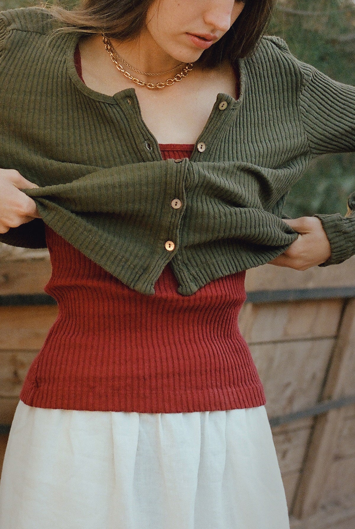 A person partially lifts the LA RELAXED Pointelle Cardigan, showing a red ribbed tank underneath. They pair it with a cream skirt and gold chain necklace. The green cardigan features pearlescent buttons; their face is mostly out of frame, photo taken outdoors.