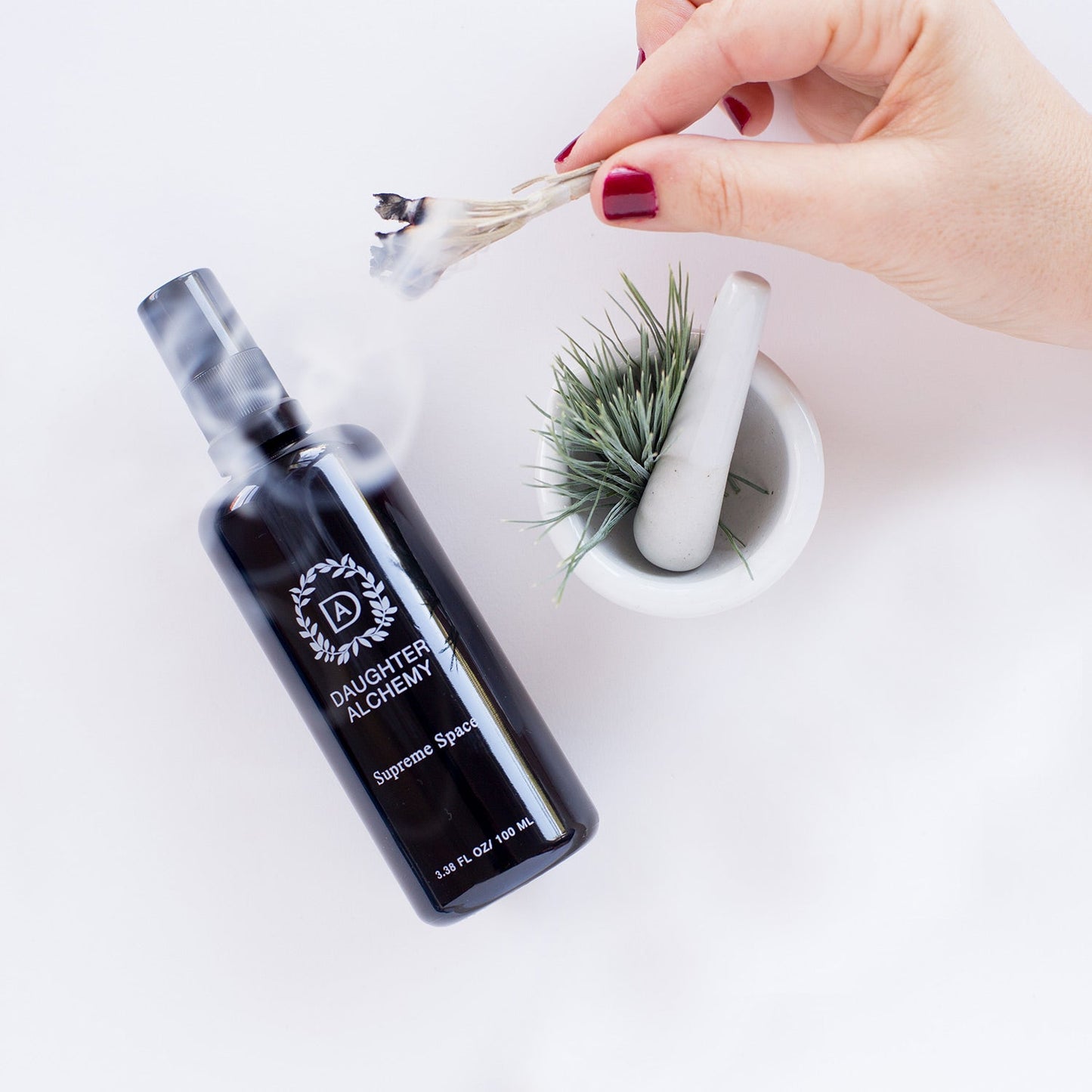 A hand with red nail polish holds a burning bundle of organic white sage above a bowl containing a small plant and a pestle. Next to the bowl is a dark bottle labeled "Daughter Alchemy - Supreme Space® Botanical Protective Spray," with smoke wafting from the bottle, balancing energies and grounding the root chakra.