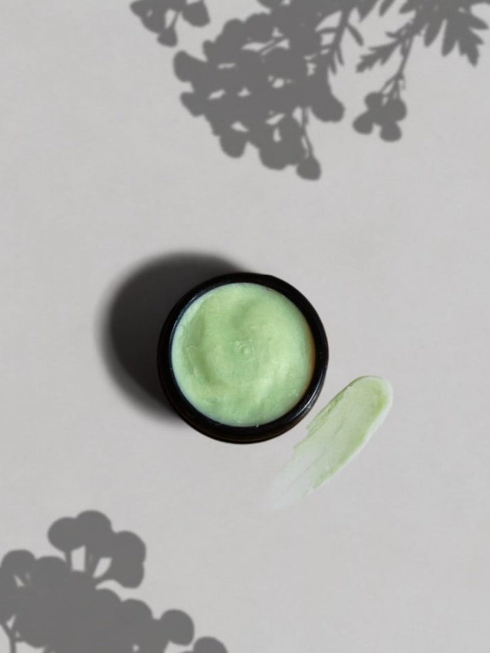 A small round jar of Wild Mamas Ancestral Blue 2.4oz grass-fed tallow cream rests on a pale surface with a smear beside it, while floral or leafy shadows in the background highlight this natural skincare moment.