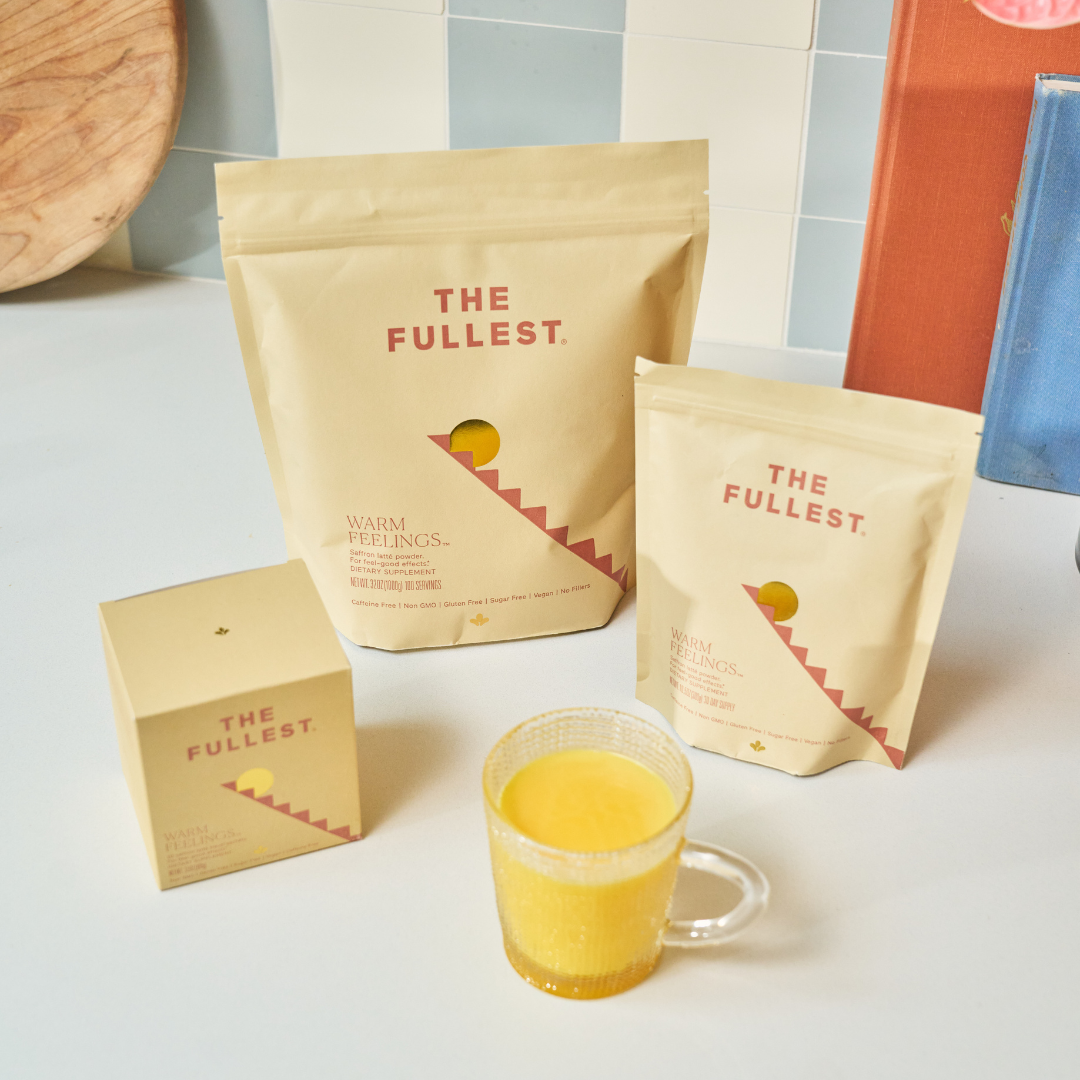 A glass mug of caffeine-free Saffron Latte 100-Day Supply by THE FULLEST sits beside three minimalist beige packages—ideal for restorative sleep and a natural mood boost, each featuring red branding and a yellow half-circle design.