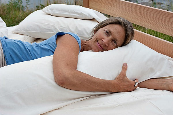 A person in a blue shirt is lying on a bed outdoors, resting their head on a Wool Filled Body Pillow from Shepherd's Dream and hugging it. The setting appears calm, with greenery in the background.
