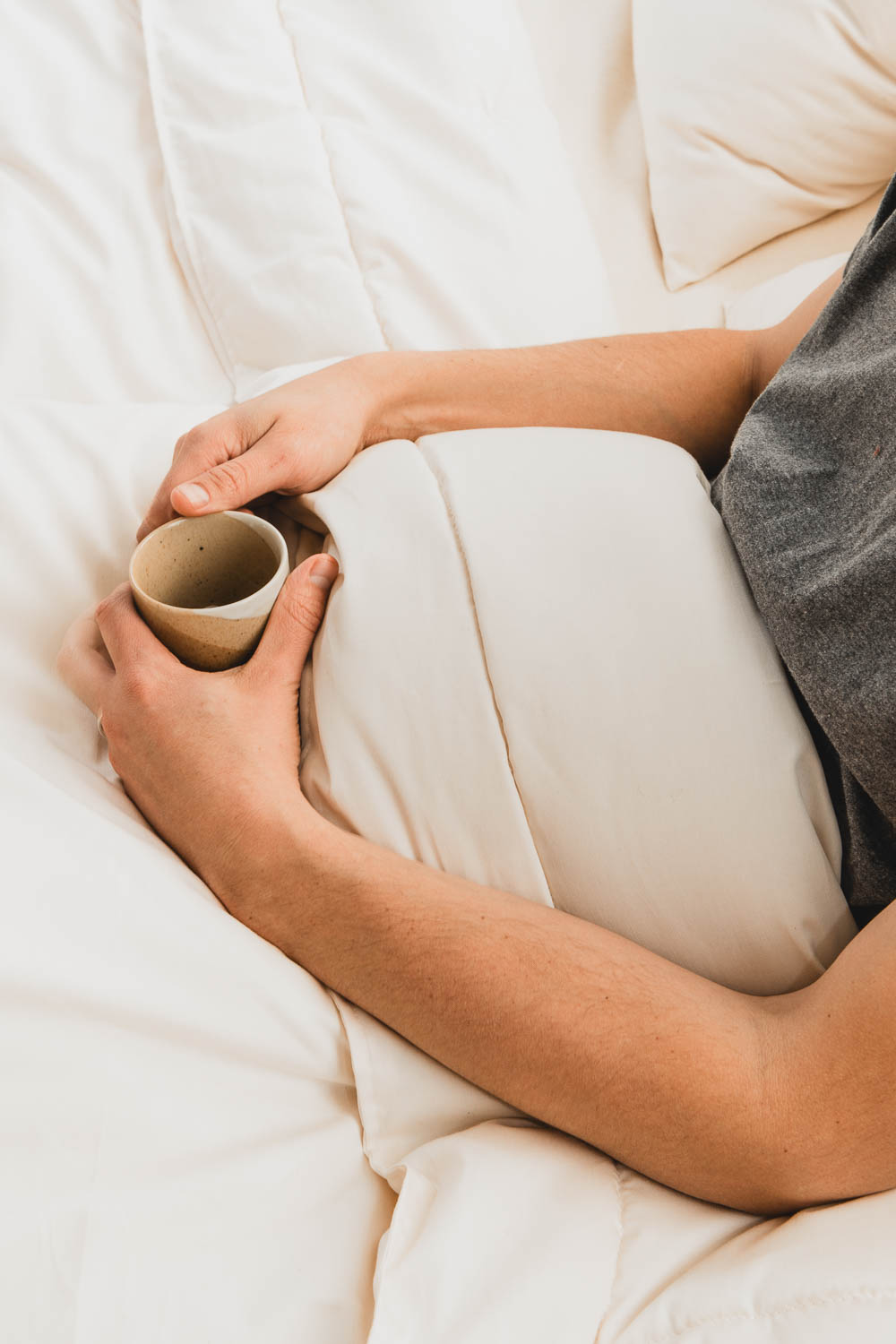 A person in a gray shirt, partially covered by the Ashland Organic Wool Comforter from Shepherd's Dream, holds a ceramic cup while sitting in bed. The scene conveys a sense of warmth and relaxation.