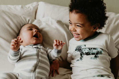 A baby in a striped onesie lies on a bed, looking up and smiling. Next to the baby, a young child wearing a dinosaur shirt sits and laughs joyfully. Both enjoy the comfort of natural bedding, surrounded by plush white *Child's Sleep Pillows* from Shepherd's Dream in the background.