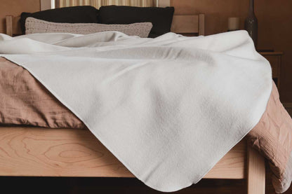 A neatly made bed features a light brown comforter and an Organic Merino Wool Blanket by Shepherd's Dream, known for its temperature-regulating properties, draped over it. Black and beige pillows are arranged at the headboard, with a nightstand and lamp visible in the background.