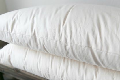 A close-up of two Ashland Organic Wool Pillows by Shepherd's Dream stacked on a wooden surface. These pillows feature a smooth texture, indicating they are clean and freshly laundered, making them ideal for a healthy bed and natural sleep environment.