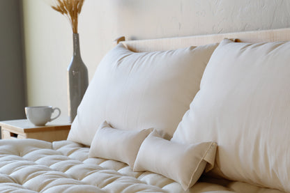 A cozy bedroom scene showcases a neatly made bed embellished with large beige pillows and a supportive Shepherd's Dream Neck Pillow for optimal cervical spine alignment. A wooden nightstand holds a white cup and a tall vase with dried grasses, creating a serene and inviting atmosphere.