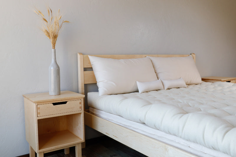 A minimalist bedroom showcases a light wood Cascade Latex Mattress Base by Shepherd's Dream, adorned with white bedding crafted from organic cotton encasement. Two pillows and small cushions lie atop the bed. A matching wooden nightstand supports a tall white vase containing dried grasses, while light-colored walls contribute to the airy ambiance.