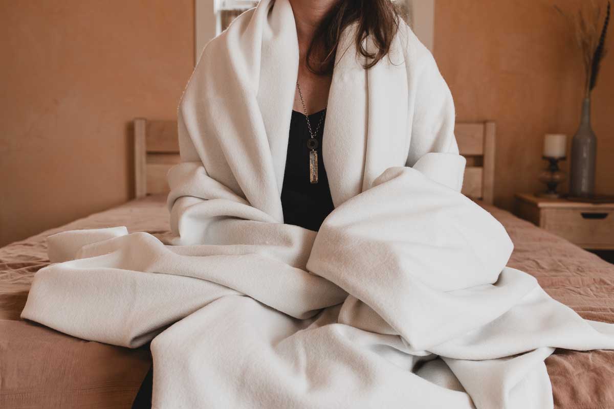 A person sits on a bed wrapped in a large Shepherd's Dream Organic Merino Wool Blanket. They are wearing a dark top and a pendant necklace. The room, with its warm-toned walls and wooden furniture, exudes cozy comfort while the temperature-regulating Merino wool ensures the perfect ambiance.