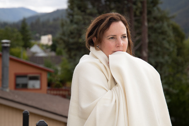 A woman wrapped in an Organic Merino Wool Blanket from Shepherd's Dream stands outdoors, looking thoughtful. Behind her, trees surround a red house set against distant hills under a cloudy sky.