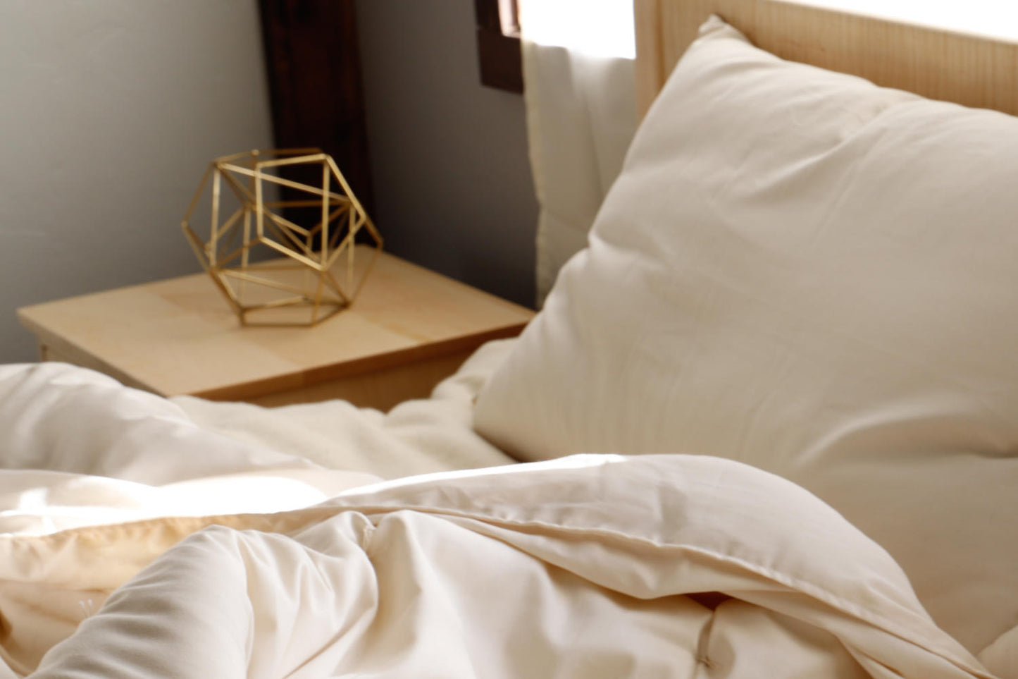 A cozy bedroom scene is complete with a neatly made bed showcasing soft, cream-colored bedding and the luxurious Ashland Organic Wool Pillow by Shepherd's Dream. A geometric gold decor piece adorns a wooden nightstand beside the bed, as sunlight streams in through a nearby window.