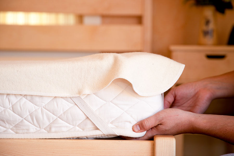 A person is putting the Shepherd's Dream Wool Mattress Protector over a white mattress. This cover adds a natural flame-resistant layer to the mattress, which features a quilted pattern and elastic strap. The setup is in a room filled with light wooden furniture and soft lighting, creating a warm and cozy atmosphere with the added assurance of safety.