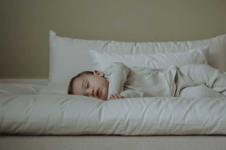 A baby sleeps peacefully on a soft, white mattress, nestled among Wool Filled Body Pillows by Shepherd's Dream. Dressed in light striped sleepwear, the child is surrounded by a serene atmosphere with a light beige background. The gentle support from the EcoWool® fill enhances the restful scene even further.
