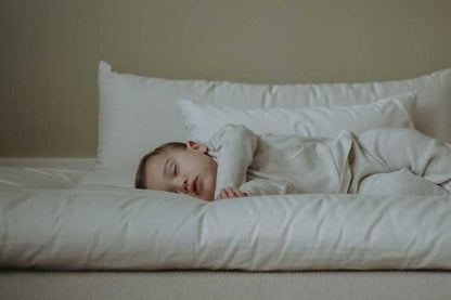 A baby sleeps peacefully on a soft, white mattress, nestled among Wool Filled Body Pillows by Shepherd's Dream. Dressed in light striped sleepwear, the child is surrounded by a serene atmosphere with a light beige background. The gentle support from the EcoWool® fill enhances the restful scene even further.